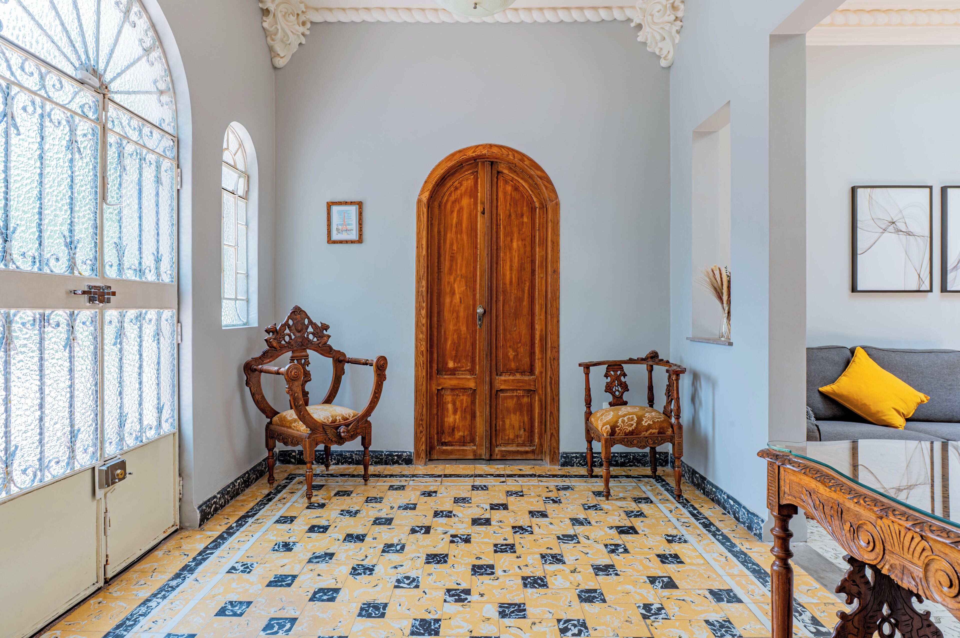 Une pièce dotée d’une grande porte en bois, de deux fauteuils richement décorés, d’un canapé avec un coussin jaune et d’un sol carrelé orné d’un motif géométrique.