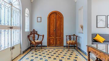 Une pièce dotée d’une grande porte en bois, de deux fauteuils richement décorés, d’un canapé avec un coussin jaune et d’un sol carrelé orné d’un motif géométrique.