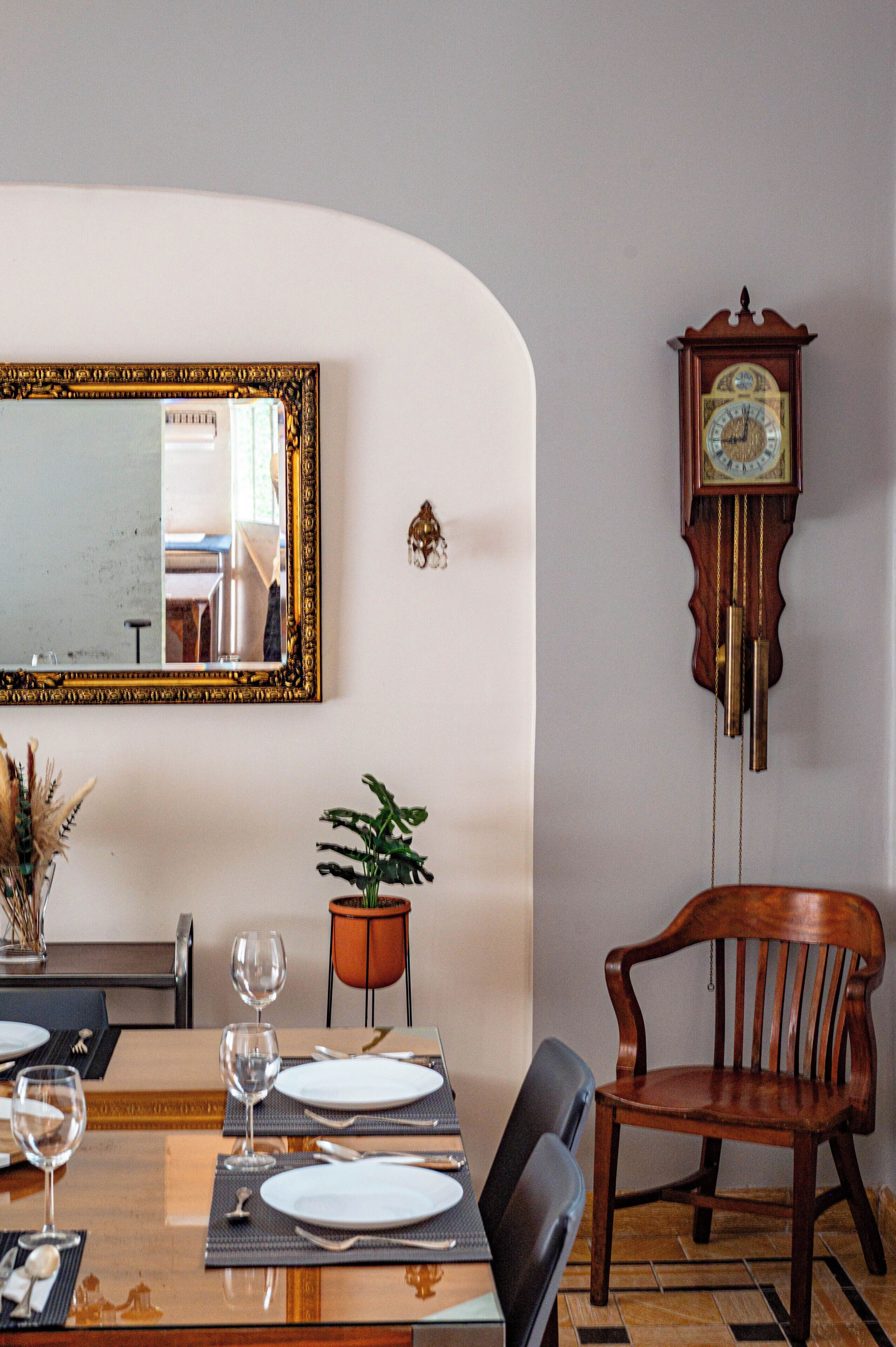 A dining area with a table set for two, a mirror, a potted plant, and a grandfather clock.