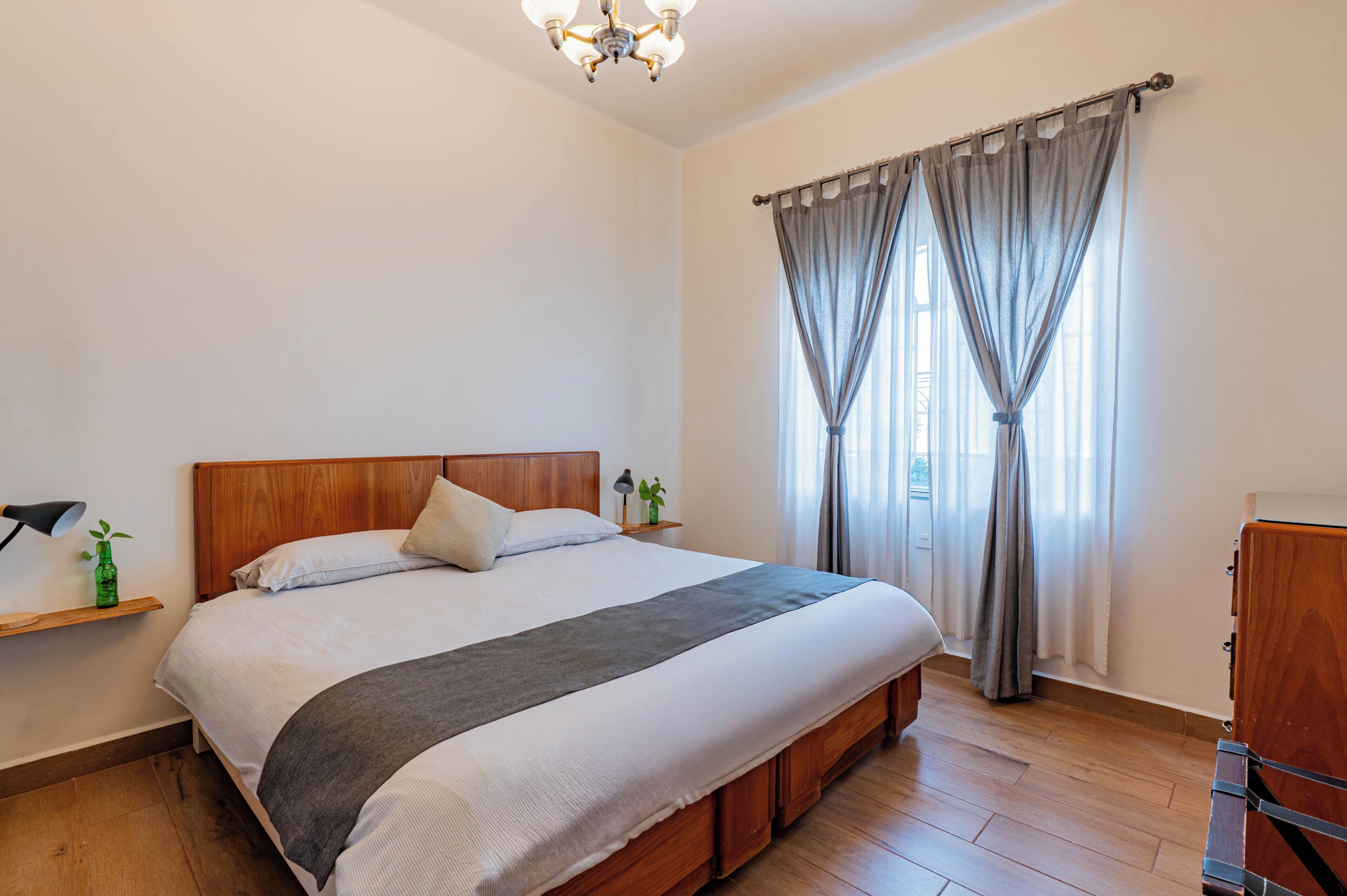 Une chambre à coucher avec un lit en bois, une table de chevet avec une lampe, une petite table de nuit avec une plante et une fenêtre avec des rideaux.