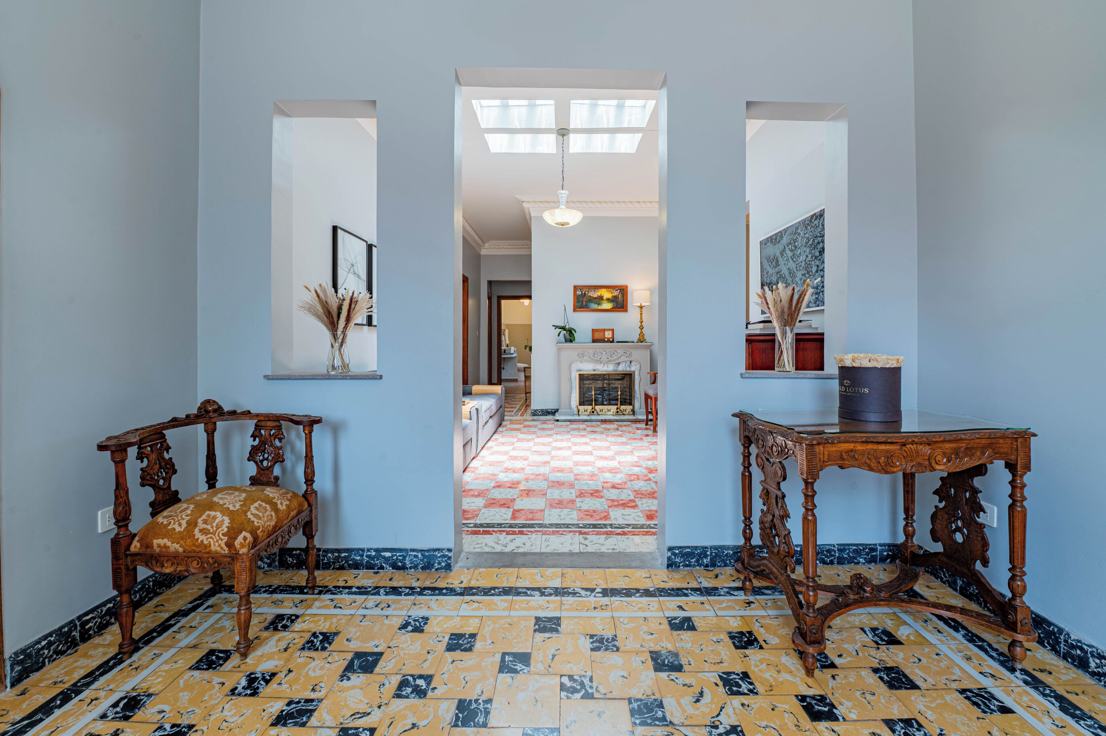 Un couloir avec un sol à carreaux, une chaise en bois, une table et une cheminée au fond.