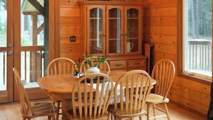 Interior de una cabaña de madera con una mesa de comedor, sillas, una araña y un aparador con puertas de cristal.
