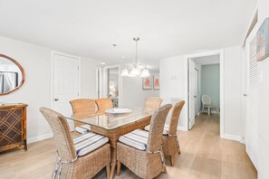 Un coin repas avec une table à plateau de verre, entourée de chaises en osier dotées de coussins à rayures, un luminaire suspendu et une porte donnant sur une autre pièce.