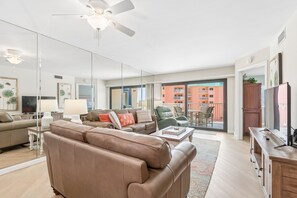 Un salon moderne avec un canapé, des fauteuils, une table basse et un téléviseur. Il y a un balcon avec vue sur un immeuble.