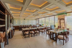 A spacious dining area with multiple tables and chairs, a kitchen in the background, and large windows offering a view of the outdoors. - Alivaa Hotel, Mcleodganj With Best Dhauladhar View (Dharamshala)