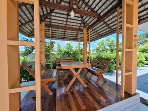 Un área de estar al aire libre cubierta, con una mesa de madera y bancos, rodeada por una estructura de madera con paneles de vidrio.