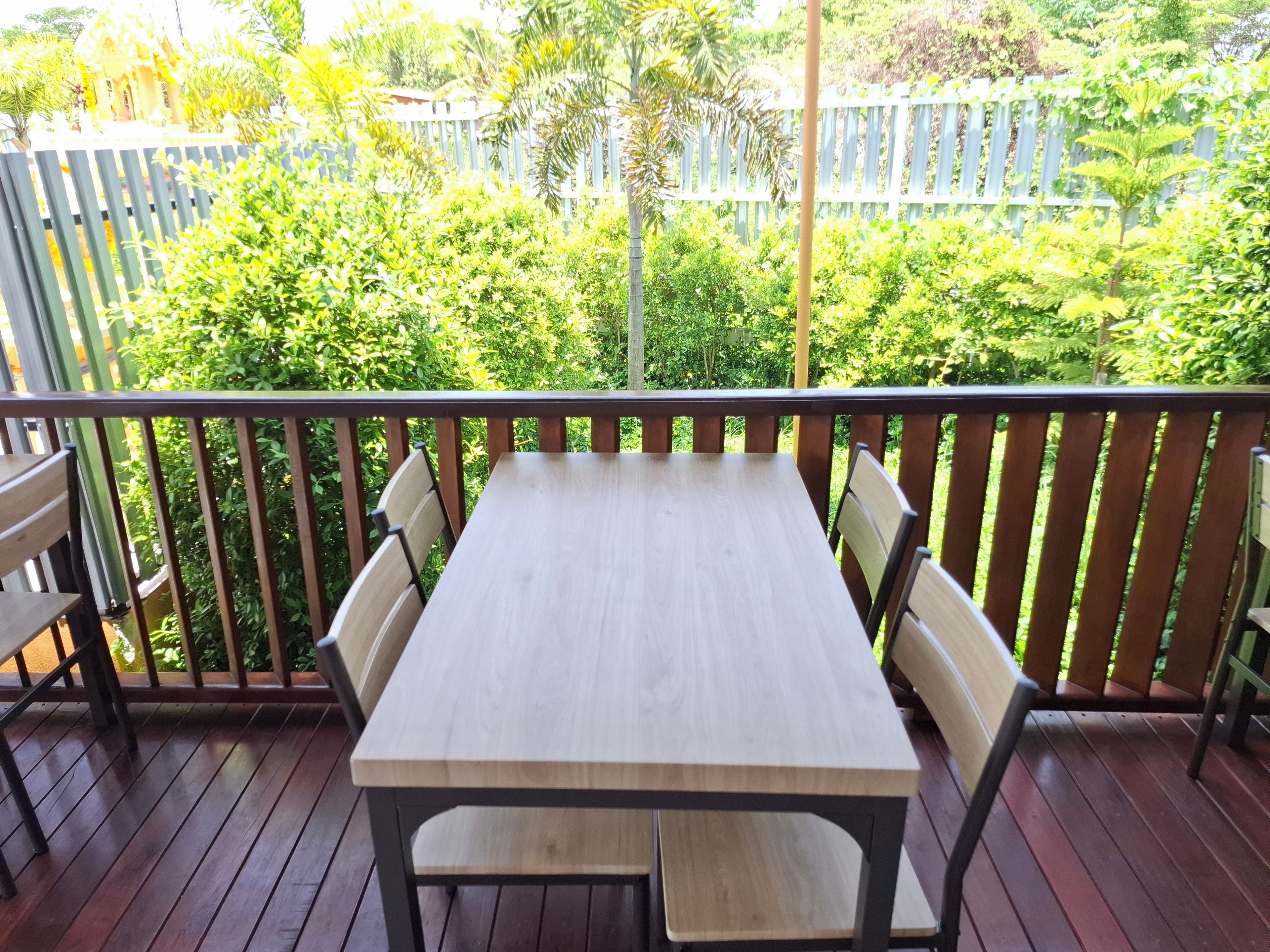 Una terraza de madera con una mesa y sillas, rodeada por una cerca blanca y abundante vegetación.
