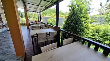 An outdoor seating area with tables and benches, a covered roof, and a view of greenery.