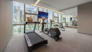 Un gimnasio moderno con caminadora, elíptica y máquinas de pesas, amplias ventanas con vista a la ciudad y una pantalla digital.