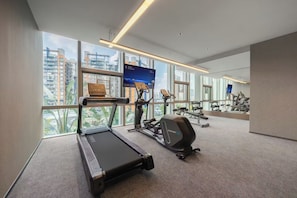 A modern gym with a treadmill, elliptical, and weight machines, large windows with a city view, and a digital display.