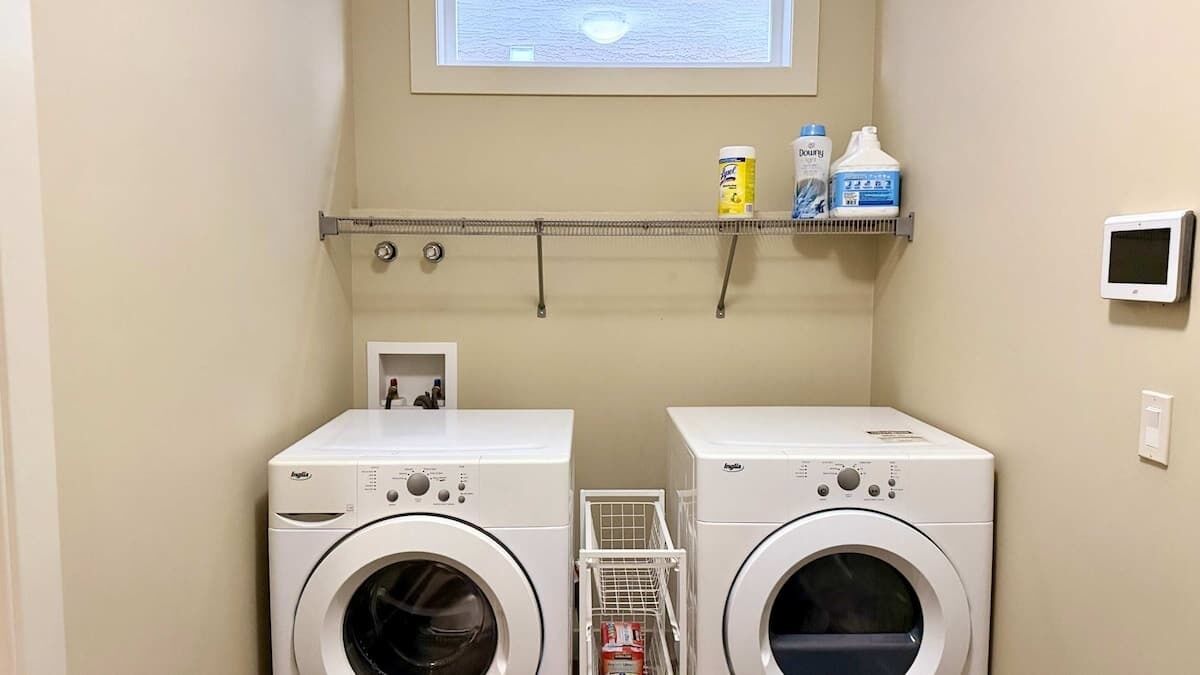 A laundry room with two front-loading washing machines, a shelf with cleaning supplies, and a small window above the machines.