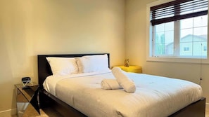 A neatly made bed with white linens and a rolled towel, a bedside table with a lamp, and a window with blinds. - Mountain View Retreat w/ Fireplace in Aspen Hills (Calgary)