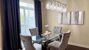 A dining area with a glass table and cushioned chairs, a chandelier, and a large abstract painting on the wall.