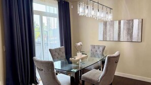A dining area with a glass table and cushioned chairs, a chandelier, and a large abstract painting on the wall.
