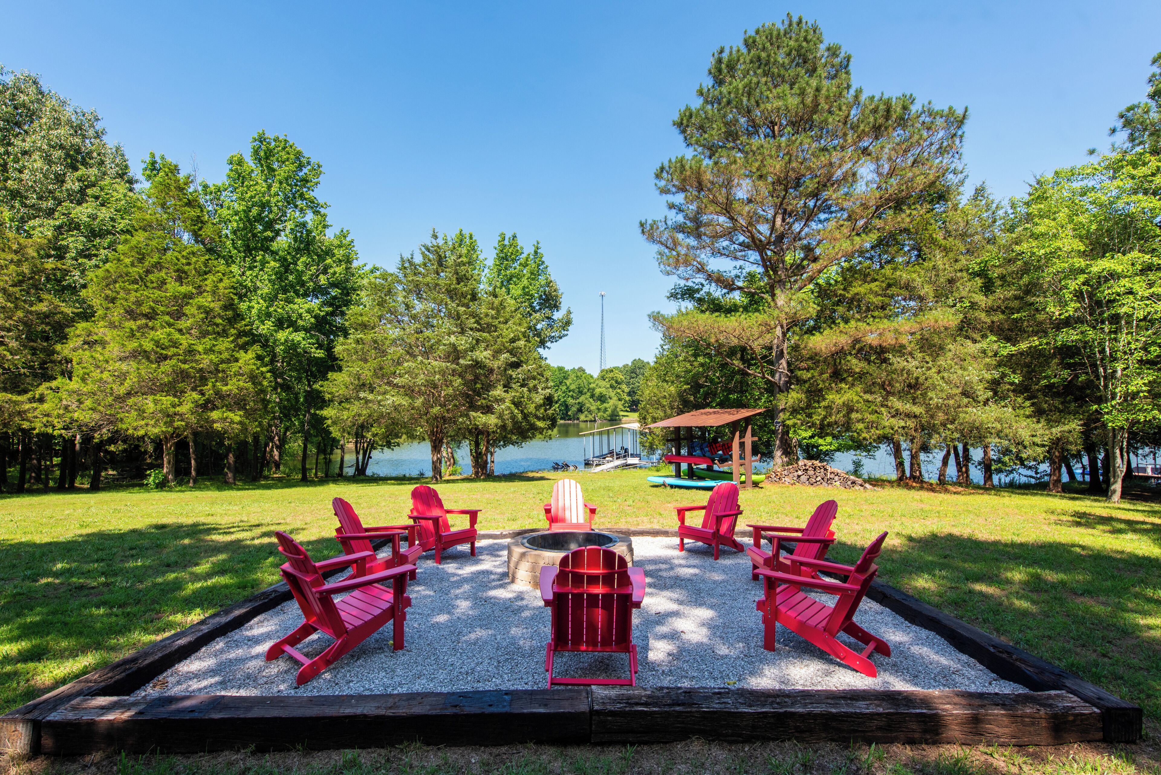 A lakeside outdoor seating area with red Adirondack chairs and a fire pit.
