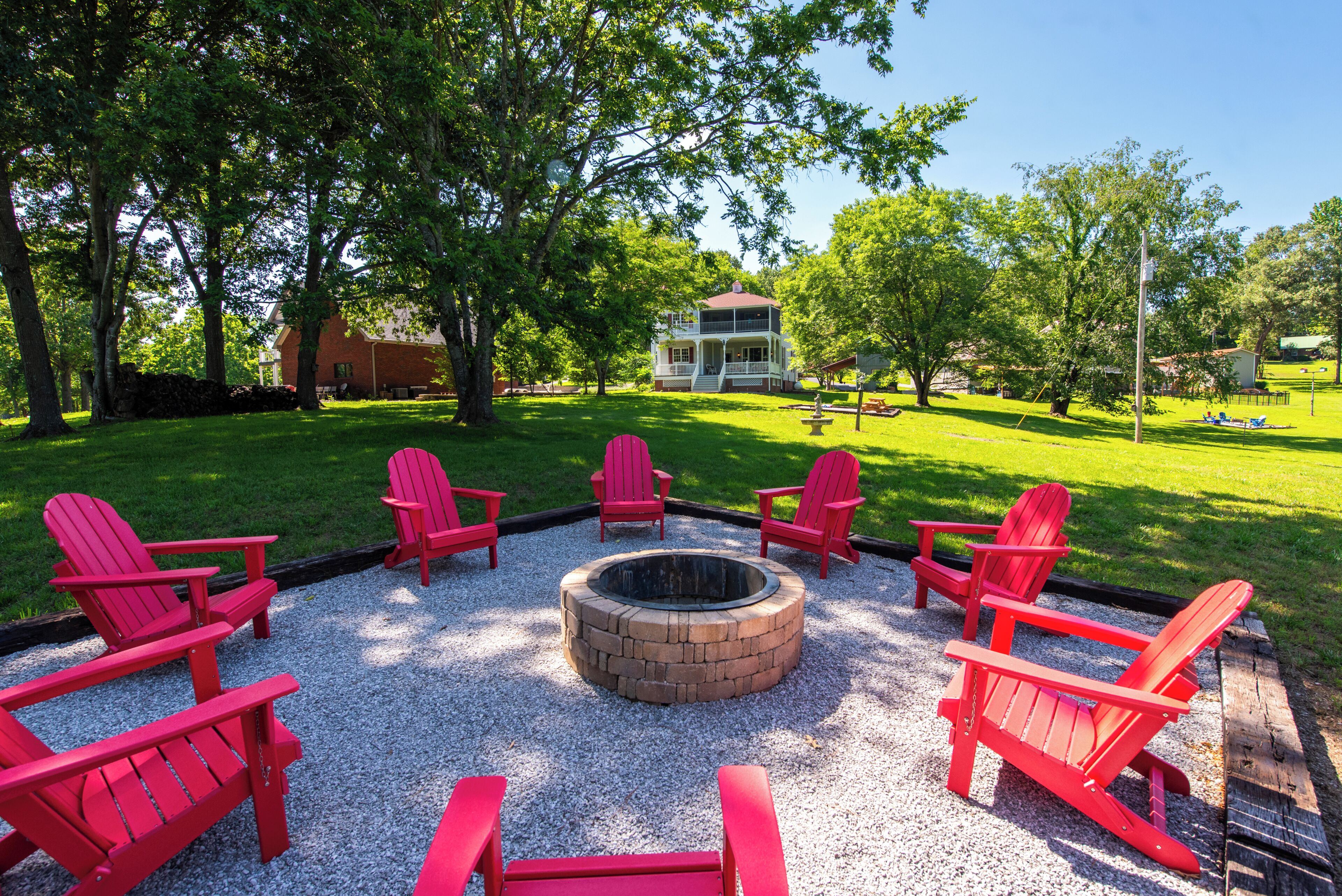 A fire pit surrounded by red Adirondack chairs in a grassy outdoor area with trees and a two-story building in the background.