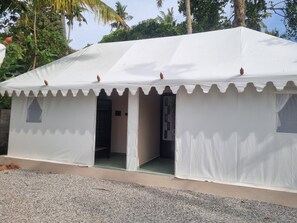 A white tent-like structure with a door and windows, surrounded by gravel and palm trees.