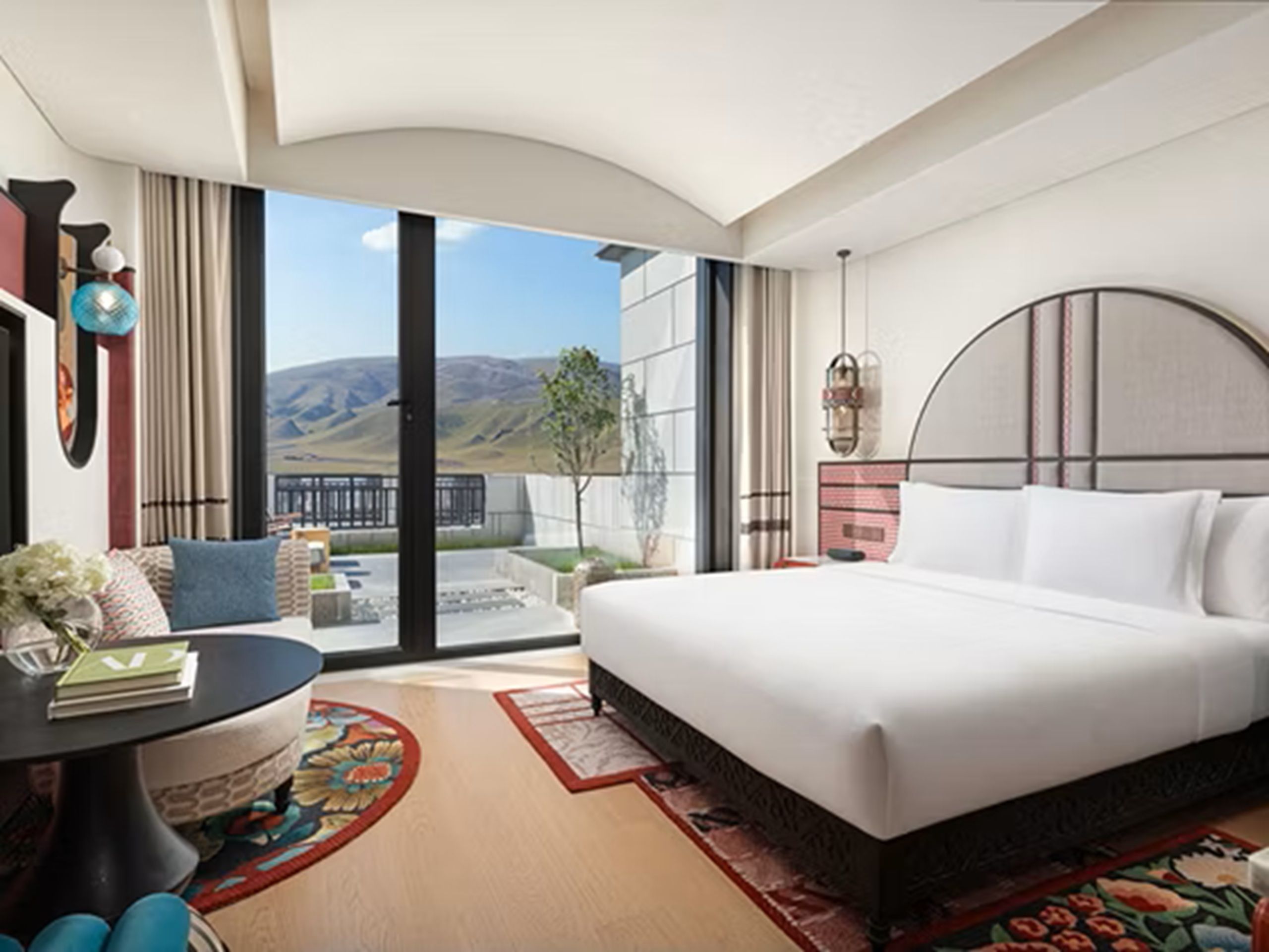 A modern hotel room with a large bed, a round table, and a view of the mountains.