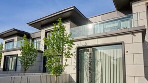 Un bâtiment moderne doté d’un toit plat, de grandes fenêtres et d’un petit espace jardin avec un banc et un arbre.