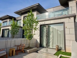 Un edificio moderno con techo plano, amplios ventanales y un pequeño jardín con un banco y un árbol.