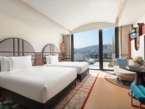 A hotel room with a large bed, a desk, a chair, and a view of the mountains.