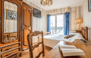 Ein Hotelzimmer mit einem Holzkoffer, einem Schreibtisch mit einem Buch, einem Bett, einem Fernseher und einem Balkon mit Vorhängen.