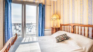 A hotel room with a bed, a nightstand, a lamp, a window with curtains, and a view of boats. - Lovely apartment in binic-étables-sur-mer (Binic-Étables-Sur-Mer)