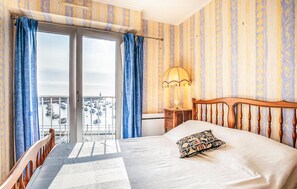 A hotel room with a bed, a nightstand, a lamp, a window with curtains, and a view of boats. - Lovely apartment in binic-étables-sur-mer (Binic-Étables-Sur-Mer)