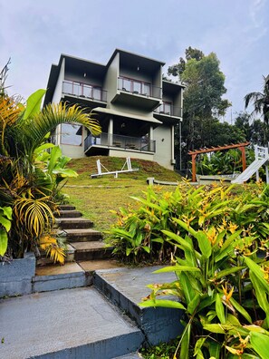A multi-story building with a balcony on the upper floor, surrounded by lush greenery and a small staircase leading to the entrance. - Studio resort in peaceful Wayanad with AC comfort (wayanad)