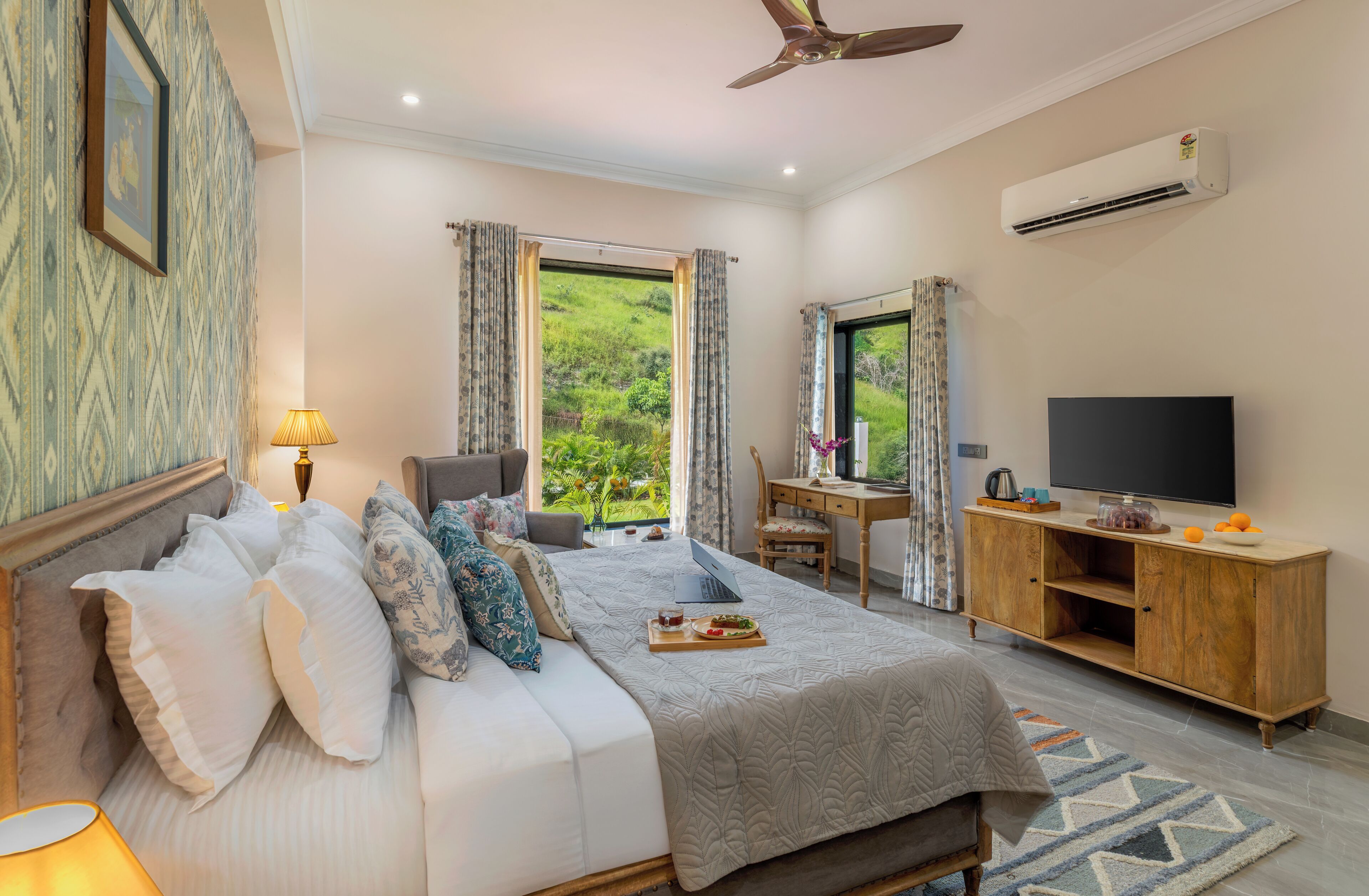A hotel room with a bed, a desk, a television, and a view of greenery.