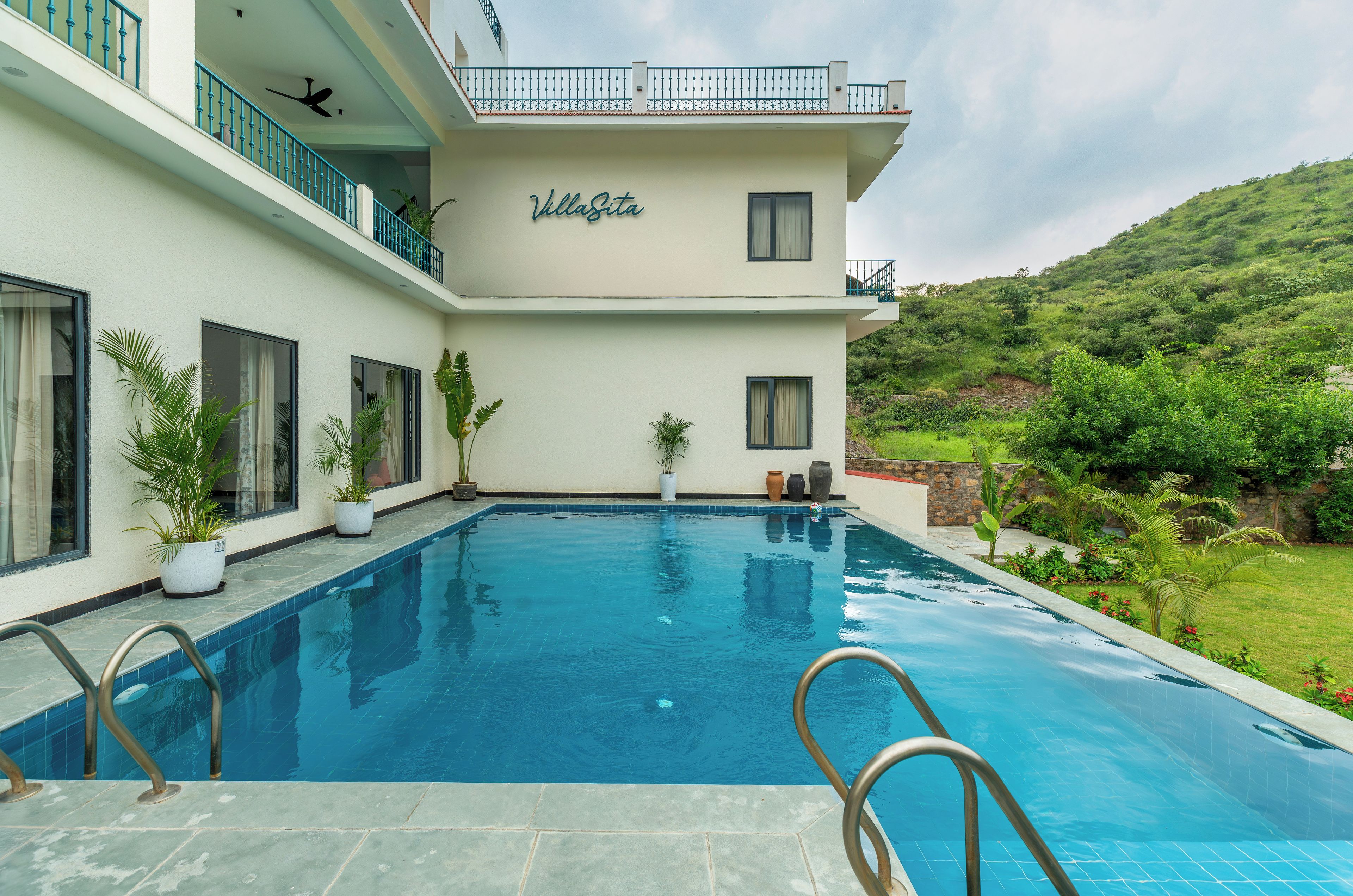 A two-story villa with a swimming pool, potted plants, and a view of a green hillside.