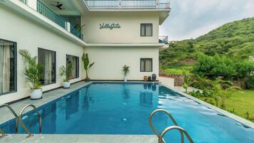 A two-story villa with a swimming pool, potted plants, and a view of a green hillside.