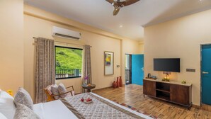 Habitación de hotel con cama, una mesa con frutas, un televisor y vista a zonas verdes.