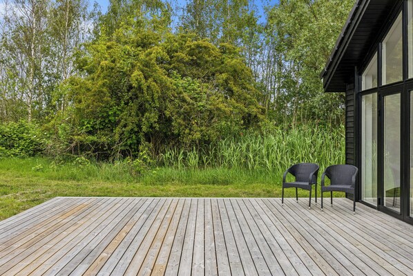 A wooden deck with two wicker chairs, surrounded by lush greenery and a large tree.
