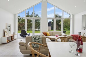 A spacious living room with a large window, a sofa, a coffee table, and a dining table with glasses and a bottle.
