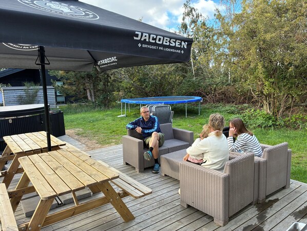 An outdoor seating area with wicker furniture, a wooden picnic table, and a black umbrella with branding.