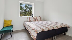 A bedroom with a bed, a window with a view of trees, a bench, and a wall-mounted light fixture. - New & inviting holiday home near beach and nature (Væggerløse)