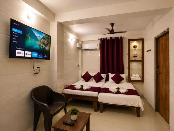 A hotel room with a flat-screen TV displaying a movie, a sofa bed with pillows, a chair, a small table with a remote, and a ceiling fan.