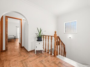 Un pasillo con suelo de madera, pared blanca, escalera de madera con barandilla y una planta en maceta sobre una mesita blanca.