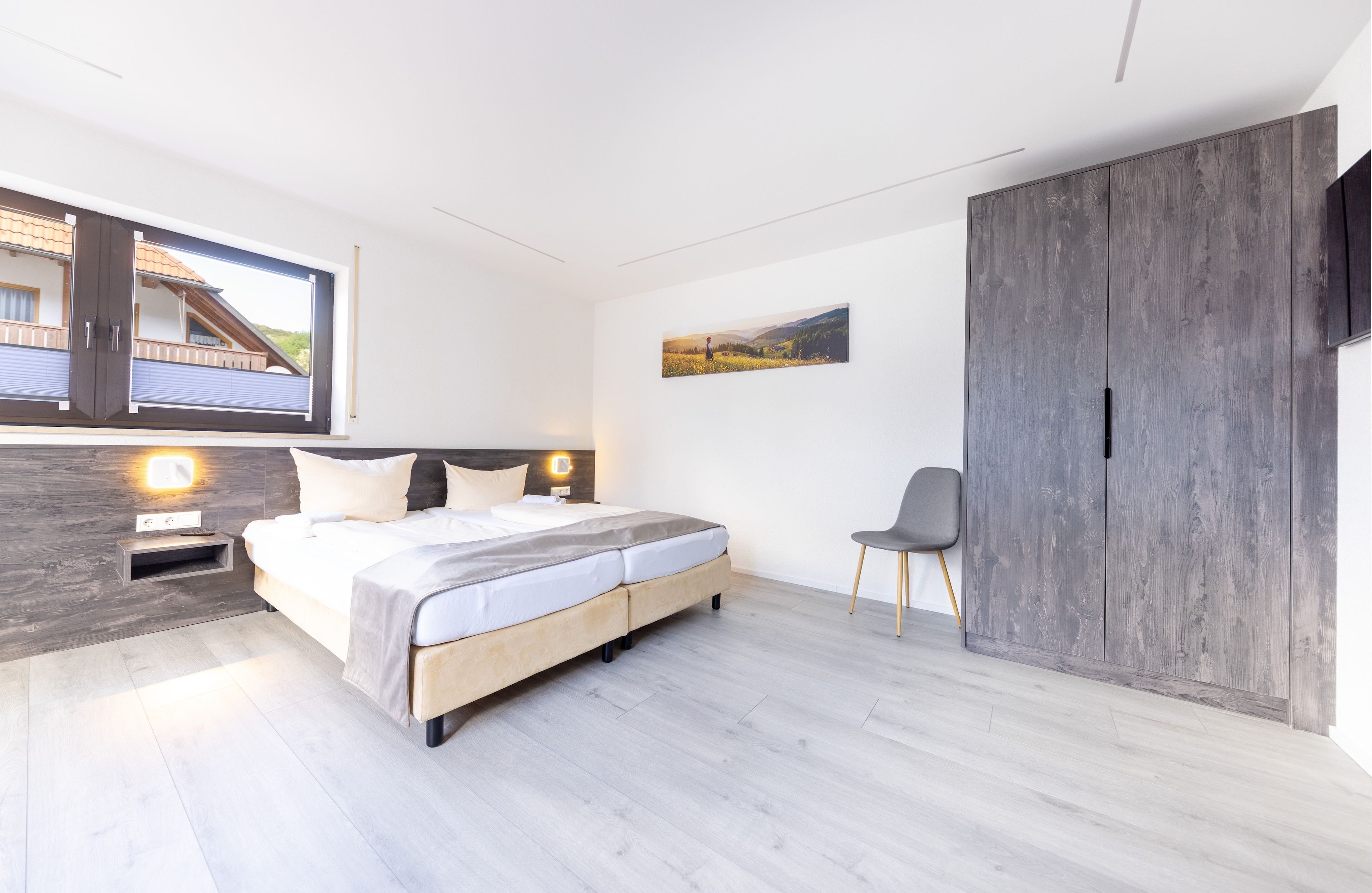 A modern bedroom with a large bed, a bedside table, a chair, a wardrobe, and a window with a view of a house.