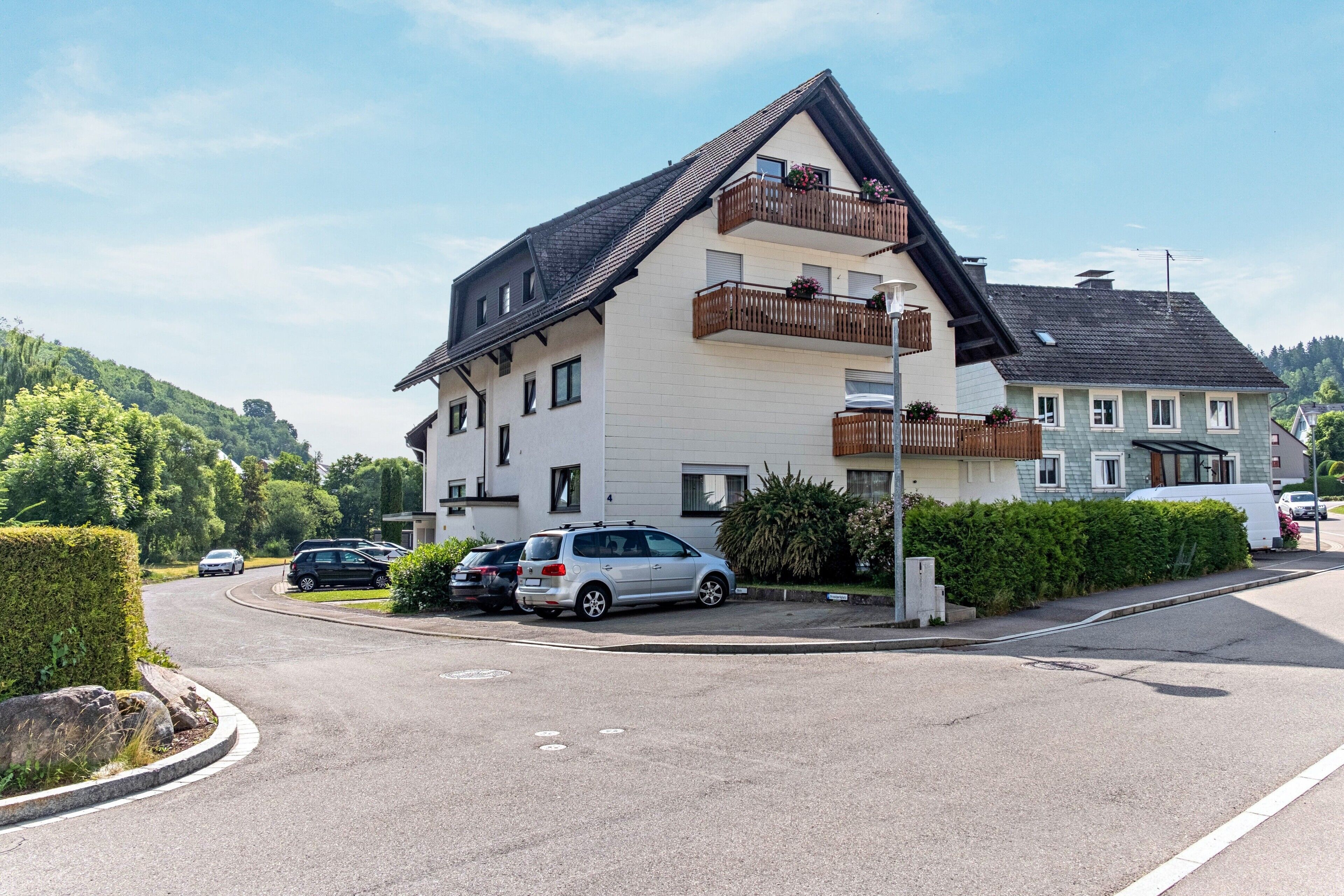 Ein Wohngebäude mit geneigtem Dach, mehreren Balkonen und einem Parkplatz mit Autos.