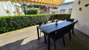 Een terras buiten met een tafel, stoelen, een geel gestreepte luifel en een haag.
