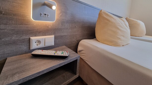 Een hotelkamer met een houten hoofdeinde, een nachtkastje met afstandsbediening en een bed met beige beddengoed.