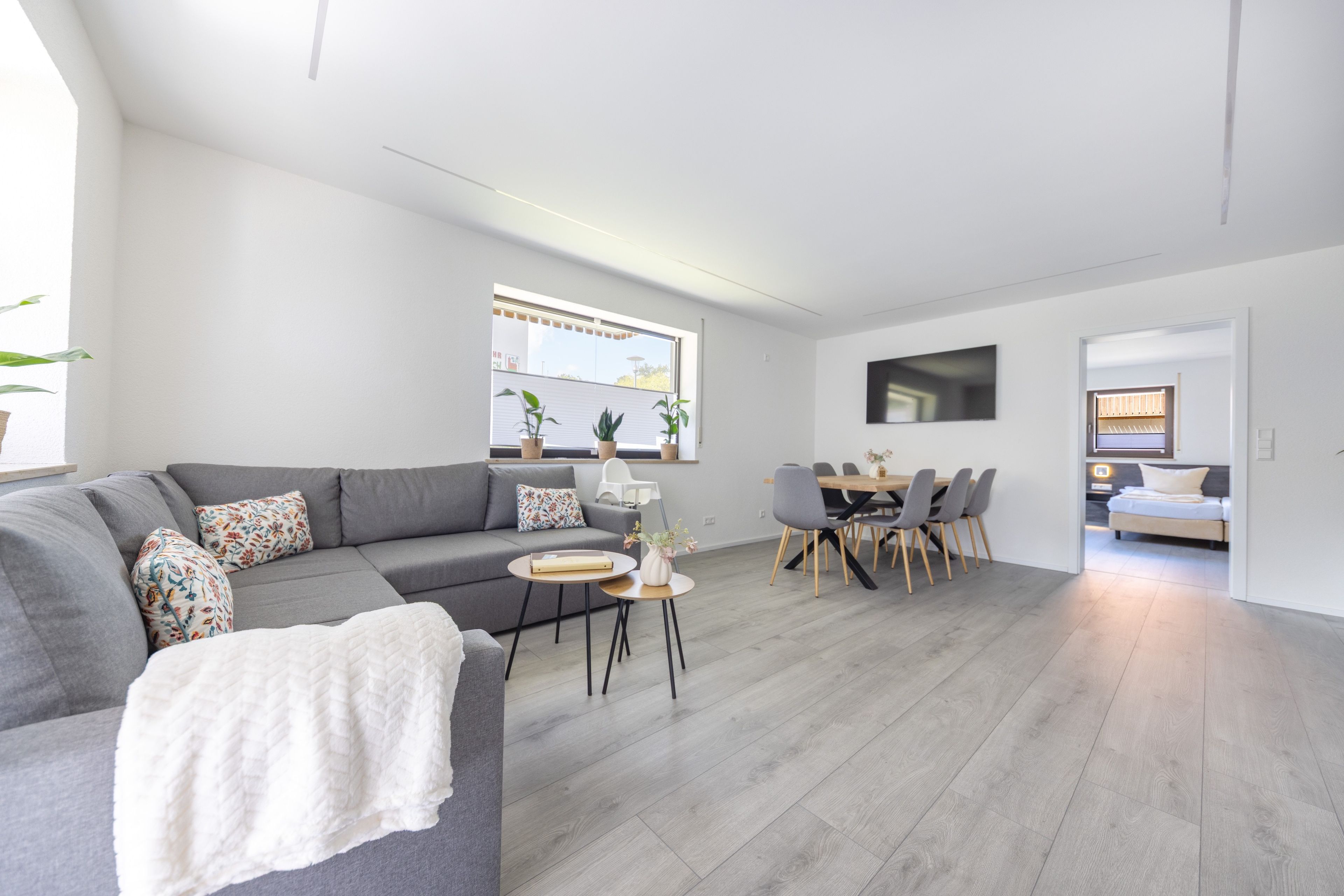 Uma sala moderna com um sofá seccional cinza, uma área de jantar com mesa e cadeiras de madeira, e um quarto com cama e televisão.
