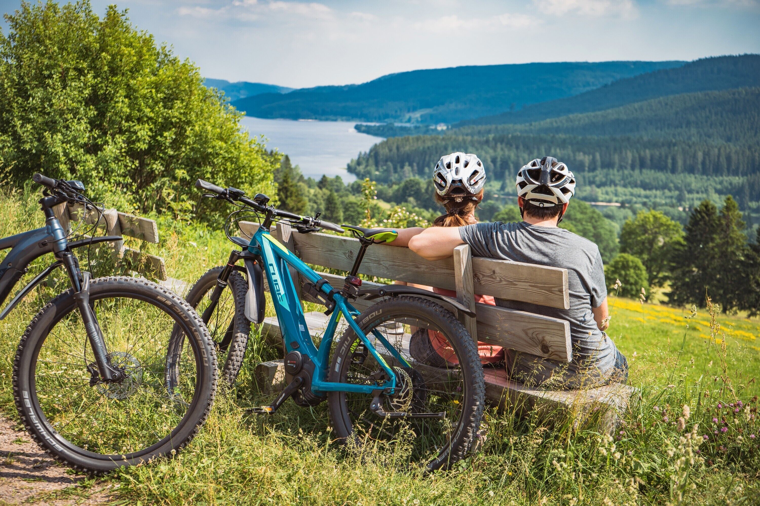 A bench with two bicycles and a person sitting on it, overlooking a scenic landscape with a lake and hills.