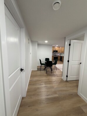 Ein Flur, der in einen Essbereich mit Tisch und Stühlen führt, im Hintergrund eine Küche und eine weiße Tür rechts.