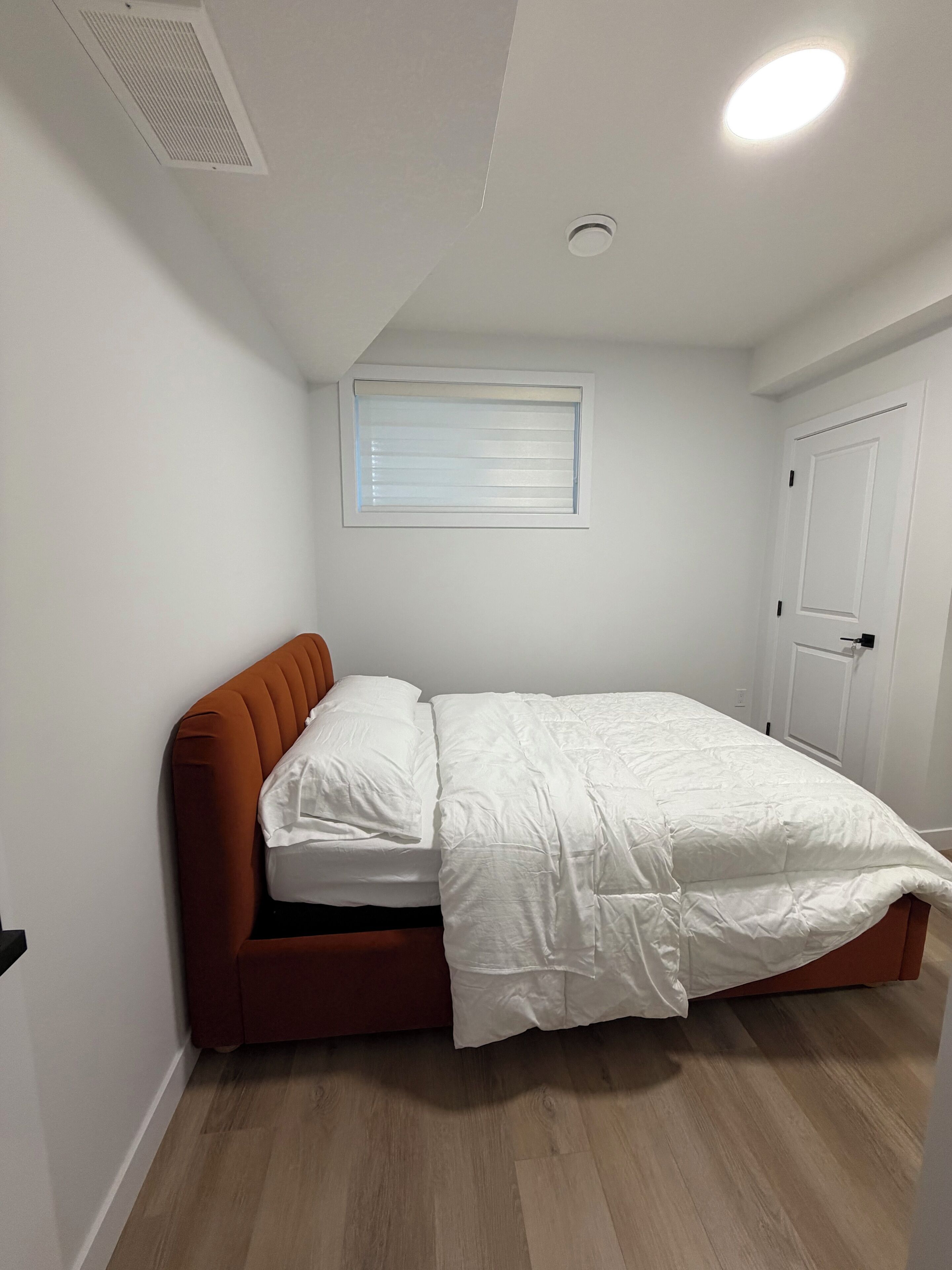 A bedroom with a single bed, a window with blinds, a door, and a ceiling vent.