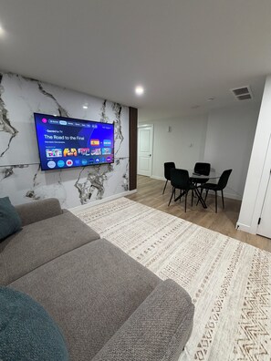 A modern living room with a flat-screen TV mounted on a marble wall, a grey sofa, a patterned rug, and a dining area with a table and chairs. - The Quarry Retreat | Modern 2-Bedroom Luxury Suite (Edmonton)