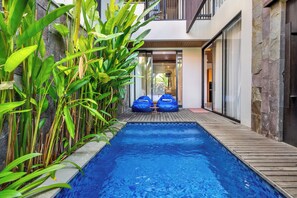 A modern outdoor pool area with a wooden deck, two blue bean bags, and lush green plants. - The Louver Canggu Exotic Bali Properties (Canggu)