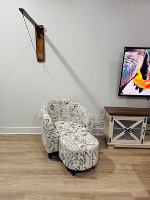 A room with a curved white armchair with black text, a wooden side table, and a television on the wall. - Cozy 1-bedroom loft apartment Downtown Dallas, gym access and nice pool (Dallas)